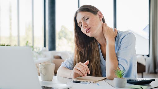 Eine Frau sitzt an einem Schreibtisch mit einem Laptop, hält ihren Nacken und sieht erschöpft aus. Auf dem Tisch liegen ein Notizbuch, ein Stift, eine Tasse und eine kleine Pflanze. Im Hintergrund sind große Fenster mit Blick auf eine verschwommene Landschaft zu sehen. Die Szene vermittelt das Gefühl von Müdigkeit oder Nackenschmerzen während der Arbeit.