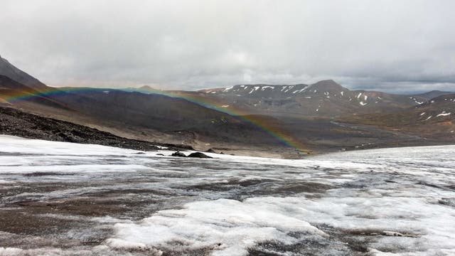 Eine Gletscherlandschaft mit schneebedecktem Vordergrund und einem Regenbogen, der sich über das Tal erstreckt. Im Hintergrund sind schroffe Berggipfel unter bewölktem Himmel zu sehen. Die Szene vermittelt eine ruhige, natürliche Schönheit.