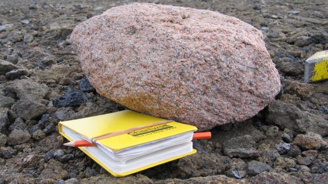 Ein großer, rötlich-brauner Felsbrocken liegt auf einer felsigen Oberfläche. Vor dem Felsbrocken befindet sich ein gelbes Notizbuch mit einem roten Stift, das auf dem Boden liegt. Im Hintergrund ist ein Gewässer zu sehen. Die Szene vermittelt einen Eindruck von Feldforschung oder geologischen Studien.