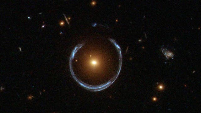 Ein Bild eines Einsteinrings, einer astronomischen Erscheinung, bei der das Licht einer entfernten Galaxie durch die Gravitationskraft einer näheren Galaxie gebogen wird, wodurch ein ringförmiges Muster entsteht. Im Zentrum des Bildes befindet sich eine helle, gelbliche Galaxie, umgeben von einem bläulichen Lichtbogen. Mehrere kleinere Lichtpunkte, die entfernte Sterne oder Galaxien darstellen, sind im Hintergrund zu sehen. Das Bild veranschaulicht das Phänomen der Gravitationslinsen in der Astronomie.