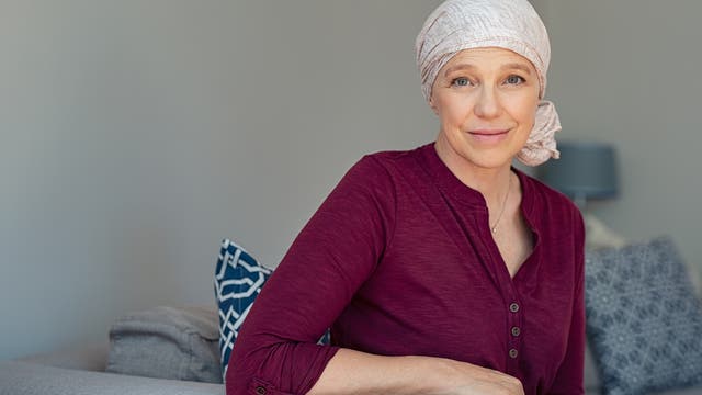 Eine Frau sitzt auf dem Sofa und hat vermutlich Krebs, da sie ein Kopftuch trägt und keine Haare hervorscheinen. Sie scheint zuversichtlich.
