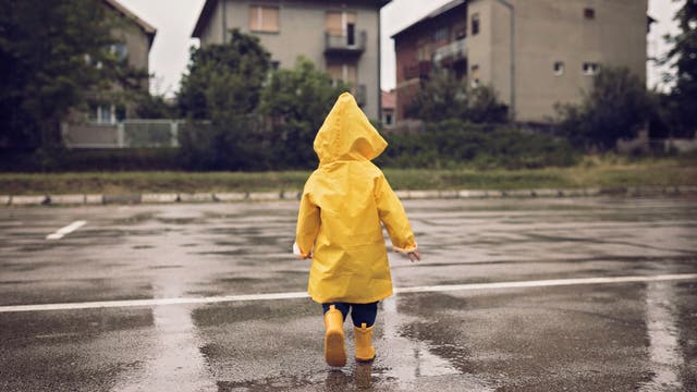 Kind in einer gelben Regenjacke und mit gelben Gummistiefeln von hinten fotografiert auf einer regennassen Straße