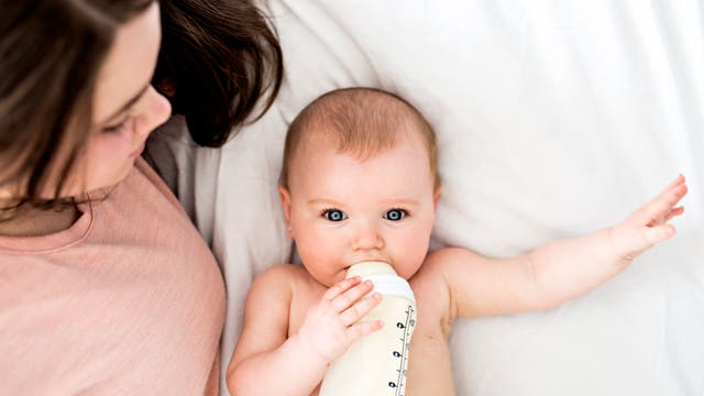 Ein Baby liegt auf dem Rücken im Bett und schaut den Betrachter direkt an, während es eine Milchflasche festhält, aus der es trinkt. Neben ihm liegt seine Mutter und schaut es an. 