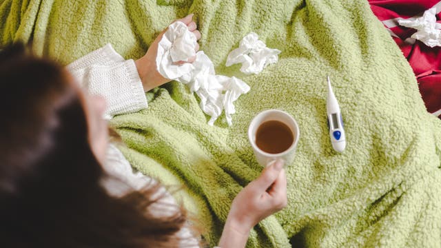 Eine Frau mit den üblichen Erkältungsutensilien: Decke, Tee, Taschentücher, Fieberthermometer.