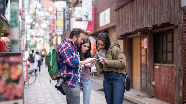 Zwei Touristen fragen eine Einheimische in den Straßen von Tokio, deren bunten Schilder man im Hintergrund sieht, mit dem Smartphone nach dem Weg.