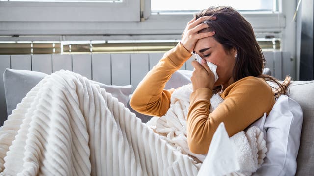 Eine Frau liegt, in eine Wolldecke gewickelt, auf ihrem Sofa. Aber sie schneuzt in ein Taschentuch, das heißt, sie ist erkältet und nicht einfach nur seit einem Jahr im Home Office.