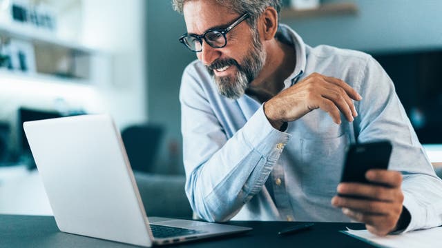 Älterer Mann mit grauem Bart und Brille schaut lächelnd in einen Laptop und hält ein Smartphone in der Hand.