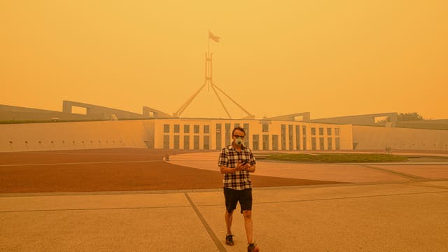 Ein Mann in Atemmaske und fragwürdig gemustertem Hemd steht vor dem australischen Parlament unter einem gelben Himmel.