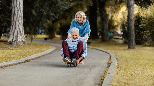 Ein grauhaariger Mann sitzt auf einem Skateboard, seine Frau schiebt ihn an. 