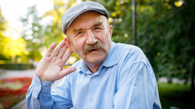 Ein älterer Mann schaut den Betrachter an und hält sich die Hand hinter das Ohr, um besser zu verstehen.