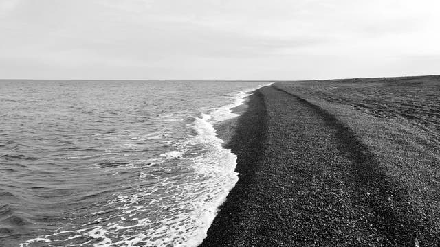 Ein schnurgerader Strandsaum aus dunklem Geröll zieht sich Richtung Horizont.