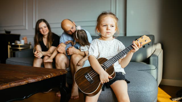 Eine Familie mit zwei Kindern sitzt im Wohnzimmer. Mutter und Vater kümmern sich um das eine Kind, während das andere Kind mit einer Gitarre auf dem Schoß traurig etwas abseits sitzt.