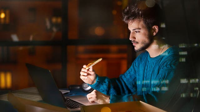 Junger mann sitzt nachts vor seinem Laptop, arbeitet und isst ein Stück Pizza.