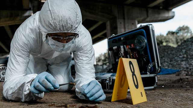 Forensiker im Schutzanzug aus Kunststoff bei der bodennahen Arbeit, vor ihm oder ihr liegen Patronenhülsen. Offenbar fand ein Schusswechsel statt. Der Tatort trägt Nummern.