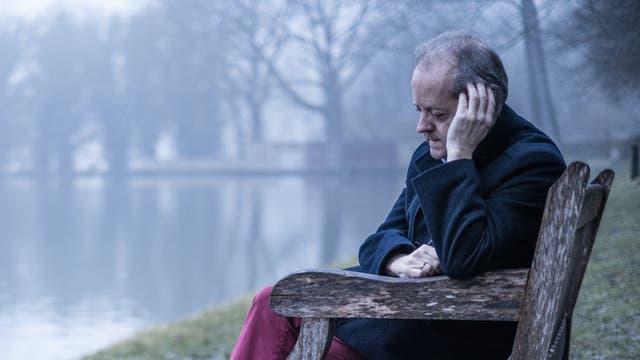 Ein Mann mit grauem Haar sitzt nach nachdenklich auf einer Bank am See 
