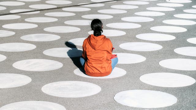 Eine Frau mit schwarzen Haaren und einer orangenen Jacke, die auf dem Boden sitzt und von hinten zu sehen ist. Auf den Boden sind weiße Punkte aufgemalt.