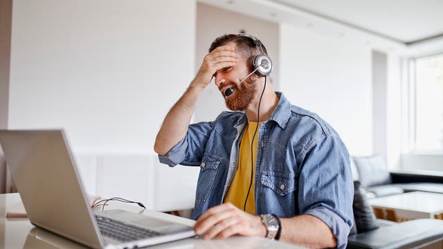 Ein Mann sitzt an einem Tisch vor einem Computer, hat ein Headset auf und fasst sich an die Stirn.