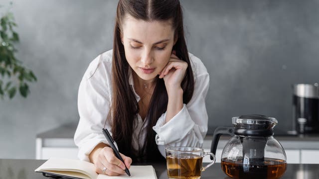 Junge Frau schreibt in ein Notizbuch, neben ihr eine Tasse und eine Kanne Tee