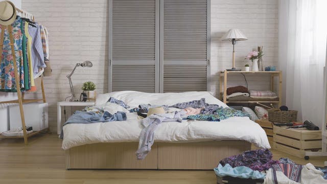 Unordentliches Schlafzimmer. Viele Kleidungsstücke liegen auf dem Bett in der Mitte des Raumes und auf dem Boden.