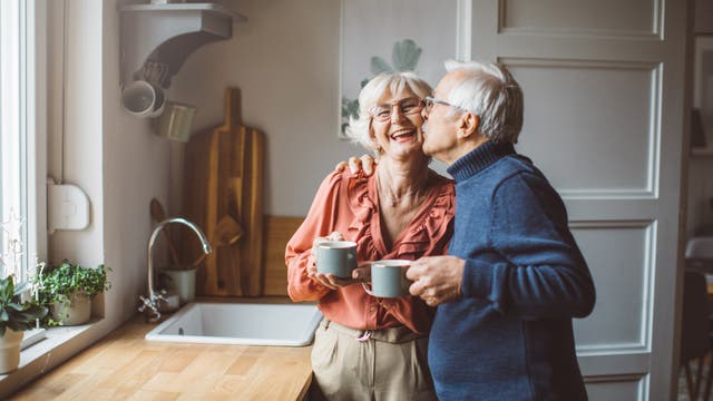Ein älteres Paar steht zusammen in der Küche mit Tassen in der Hand und der Mann gibt der Frau einen Kuss auf die Wange.