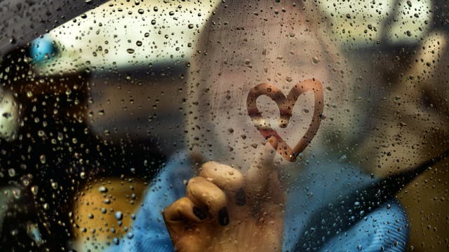 Herz, das mit dem Finger auf eine verregnete Autoscheibe gemalt wurde. Im Hintergrund ist verschwommen die Frau zu erkennen, die das Herz gemalt hat. 