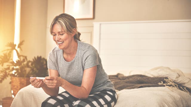 Eine Frau zwischen 30 und 40 sitzt auf dem Bett und schaut glücklich auf einen Schwangerschaftstest