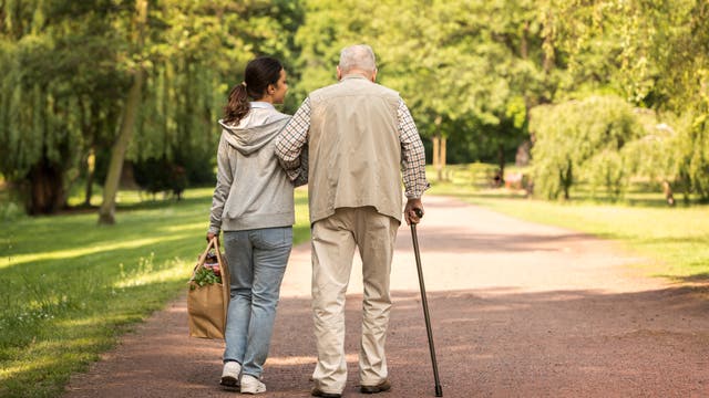 Eine junge Frau und ein älterer Mann gehen gemeinsam auf einem Parkweg spazieren. Die Frau trägt eine Einkaufstasche und hält den Arm des Mannes, der einen Gehstock benutzt. Umgeben von grünen Bäumen und einem sonnigen Himmel, vermittelt die Szene ein Gefühl von Unterstützung und Gemeinschaft.
