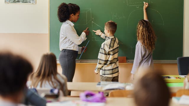 Lehrerin und zwei Kinder stehen an der Tafel, im Vordergrund sitzen andere Kinder in den Schulbänken