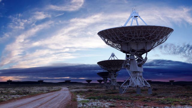 Eine Reihe von großen Radioteleskopen steht in einer offenen Landschaft unter einem dramatischen Himmel mit Wolkenformationen. Die Teleskopschüsseln sind auf Metallstrukturen montiert und richten sich gen Himmel. Ein unbefestigter Weg führt durch die Szene, die von einer weiten, flachen Landschaft umgeben ist. Die Abenddämmerung erzeugt ein sanftes Lichtspiel am Horizont.