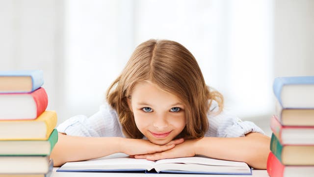 Ein Mädchen sitzt an einem Tisch und stützt sich mit den Händen auf einem Buch ab. Sie ist von Bücherstapeln umgeben.