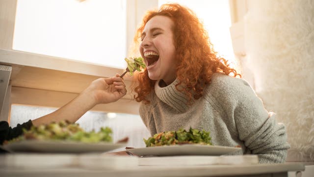 Eine jungen Frau mit roten Haaren wird von einem Mann mit Salat gefüttert. Sie lacht.