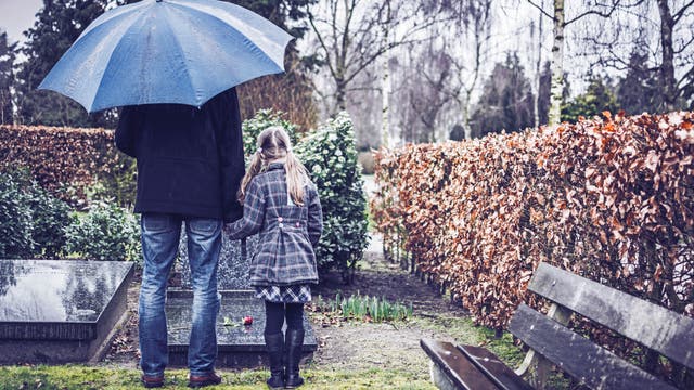 Ein Erwachsener und ein Kind stehen im Regen vor einem Grab mit Rose auf der Grabplatte..
