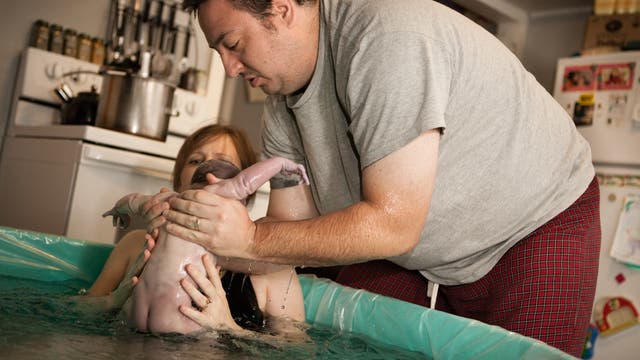Eine Frau sitzt im Wasser und hält, unterstützt vom Vater, ihr Neugeborenes.