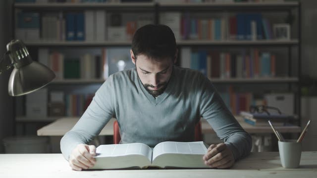 Ein Mann blickt konzentriert in ein dickes Buch. Hinter ihm steht ein Bücherregal. Eine Schreibtischlampe beleuchtet das Buch.