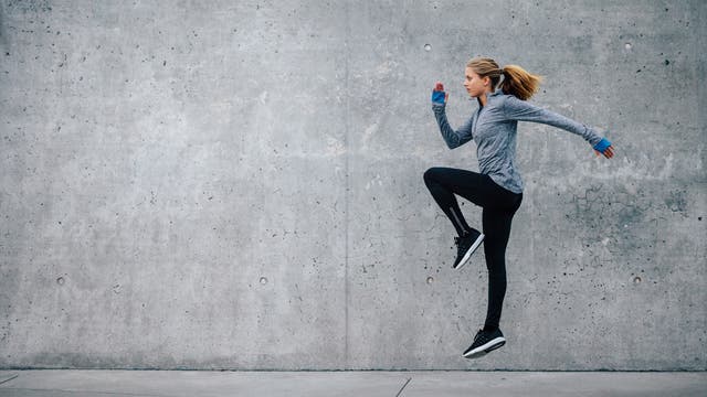 Eine Person in Sportkleidung springt dynamisch vor einer grauen Betonwand. Sie trägt eine graue Jacke, schwarze Leggings und schwarze Sportschuhe. Die Haare sind zu einem Pferdeschwanz gebunden. Die Szene vermittelt Energie und Bewegung.
