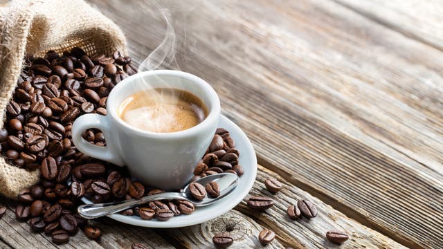 Eine Tasse Kaffee auf einem Holztisch, umgeben von Kaffeebohnen, die aus einem Jutebeutel gekippt sind. Es ist quasi das Symbolbild für Kaffee-Symbolbilder. 