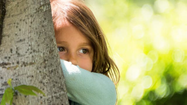 Ein kleines Mädchen steht hinter einem Baum und guckt etwas traurig