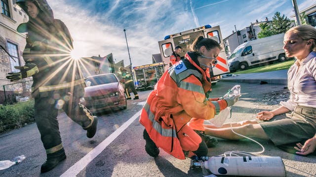 Eine Notfallärztin hilft einer bei einem Autounfall verletzten Frau, die auf einer Straße sitzt und erschöpft die Augen schließt. Im Hintergrund ist das kaputte Auto zu sehen sowie ein Rettungswagen und weitere Ersthelfer.