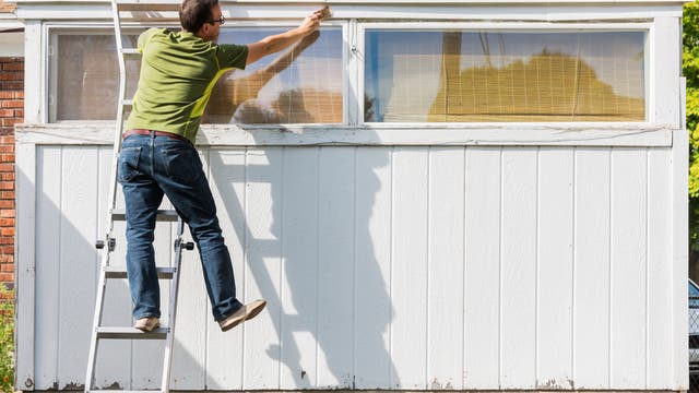 Ein Mann mittleren Alters steht auf der Leiter, streicht das Holz über einem Fenster und fällt fast von der Leiter