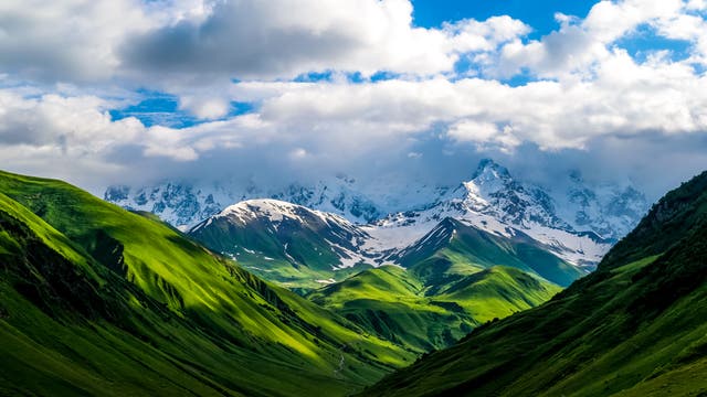 Grüne, sanfte Hügel erstrecken sich im Vordergrund, während schneebedeckte Berge im Hintergrund unter einem bewölkten Himmel aufragen. Natur im Kaukasus – die verschiedenen Flächen haben eine unterschiedliche Albedo.