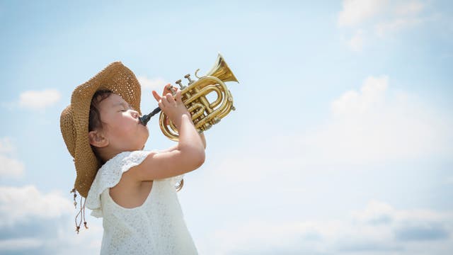 Ein kleines Mädchen mit weißem Kleid steht vor einem blauen Himmel und bläst in eine kleine Trompete.