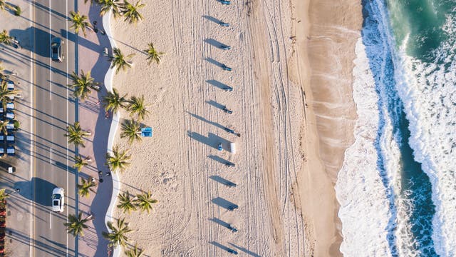 Das Bild zeigt einen Strand, an den rechts Wellen branden, während links eine Straße entlang läuft. Am Sandstrand selbst finden sich Rillen und Spuren im Sand.