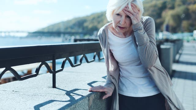 Eine ältere Frau geht draußen in der Sonne spazieren und hat Kopfschmerzen oder Schwindel. Sie fasst sich mit einer Hand an den Kopf.