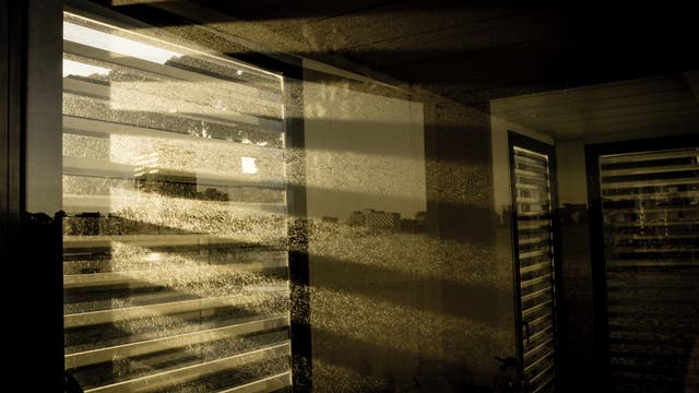 Licht fällt durch ein teilweise abgedecktes Fenster ins Innere eines Gebäudes und erzeugt ein Streifenmuster auf dem Staub des Zimmers.