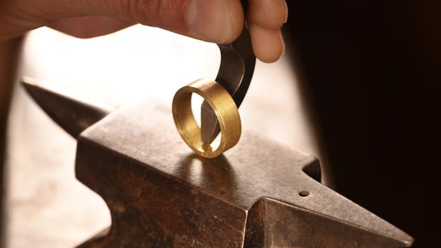 Auf einem sehr kleinen Amboss prägt ein Juwelier mit einem Schlageisen sein Logo in die Innenseite eines breiten und etwas geschmacklosen Goldringes.