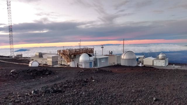 Gebäude des Mauna Loa Observatory stehen auf dem Vulkan Mauna Loa über einem Wolkenmeer. Im Hintergrund ist ein kleinerer Gipfel dunkel erkennbar. Es herrscht Morgendämmerung. Die Gebäude sind niedrig und weiß, zahlreiche Antennen sind erkennbar.