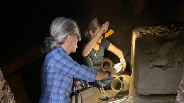 Zwei Personen arbeiten in einer archäologischen Ausgrabungsstätte. Sie befinden sich in einem dunklen Raum, der von einer Taschenlampe beleuchtet wird. Im Hintergrund sind antike Keramiken und Steine zu sehen. Eine Person hält ein Werkzeug, während die andere etwas untersucht. Die Szene vermittelt den Eindruck von konzentrierter Forschung und Entdeckung.