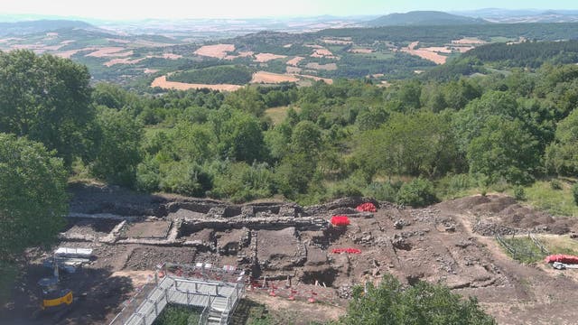 Luftaufnahme einer archäologischen Ausgrabungsstätte in einer hügeligen Landschaft. Im Vordergrund sind die Überreste von Steinmauern und rechteckigen Strukturen zu sehen, umgeben von Bäumen und Vegetation. Ein kleiner Bagger und rote Markierungen sind auf dem Gelände sichtbar. Im Hintergrund erstreckt sich eine weite Landschaft mit Feldern und Wäldern bis zu den entfernten Hügeln am Horizont.