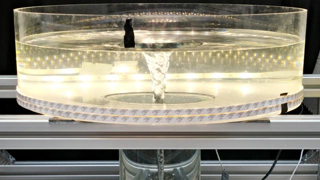 Ein runder, transparenter Wassertank mit einem sichtbaren Wasserwirbel in der Mitte. Der Tank ist von einer LED-Lichtleiste umgeben, die für gleichmäßige Beleuchtung sorgt. Der Wirbel erstreckt sich von der Wasseroberfläche bis zum Boden des Tanks. Der Aufbau befindet sich auf einer Metallstruktur.