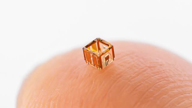 Ein winziger, transparenter Würfel mit goldenen Strukturen ruht auf der Spitze eines Fingers. Der Würfel zeigt feine, elektronische Details und ist so klein, dass er die Textur der Haut betont. Der Hintergrund ist weiß und minimalistisch, was den Fokus auf den Würfel lenkt.